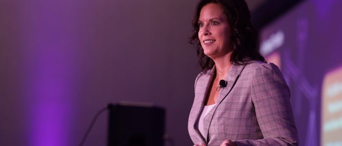 Speaker on stage before a purple-lit screen at the 2025 symposium